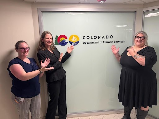Decorative Photo of Colorado CDDHHDB Team Few of Colorado Division for the Deaf, Hard of Hearing, and DeafBlind team members standing together in an indoor hallway with Colorado Department of Human Services logo on office window panels.